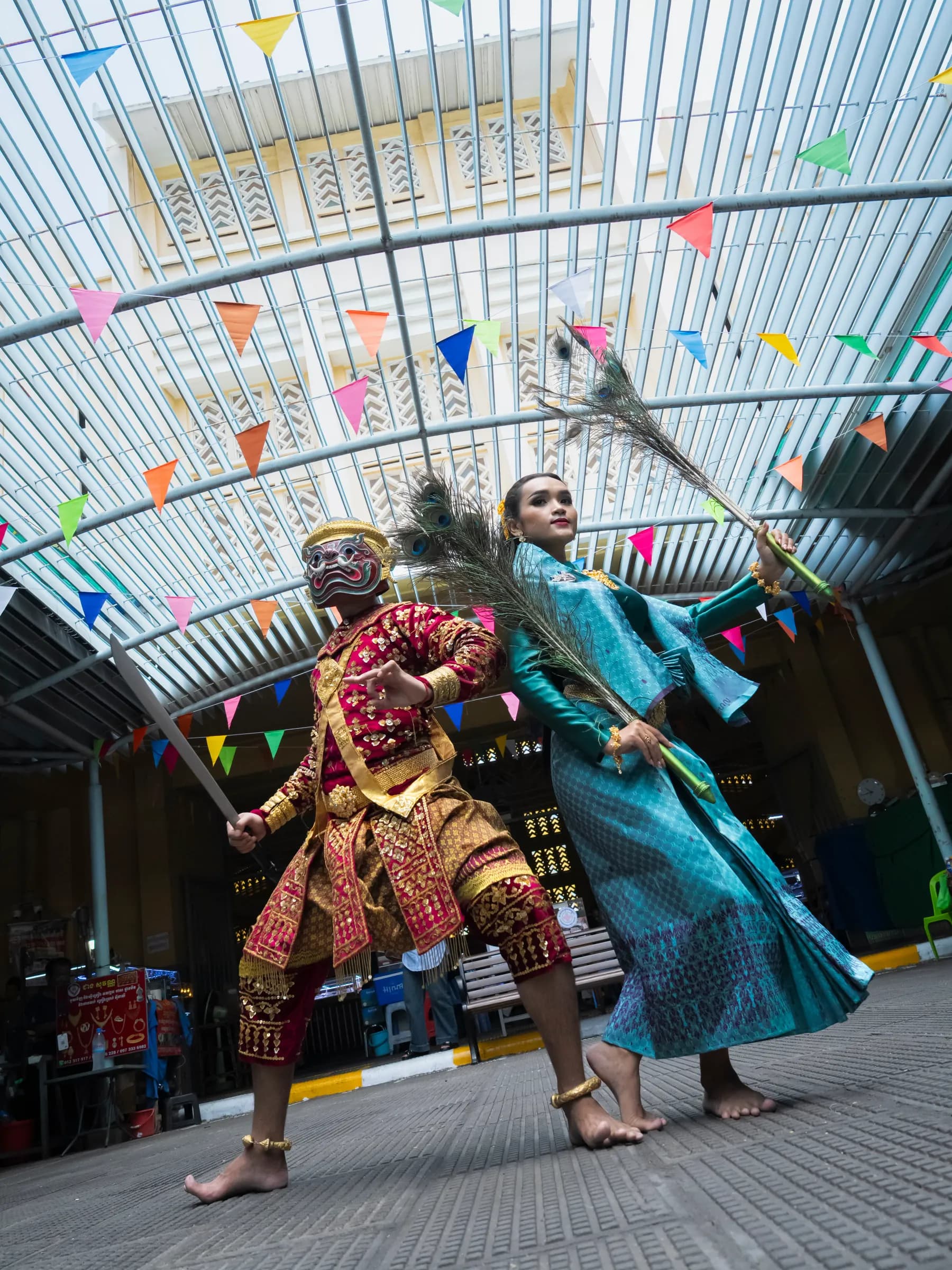 Two Robam Trot dancers at Phnom Penh’s Central Market: a masked giant in red and gold holding a sword, and a woman in teal silk holding peacock feathers