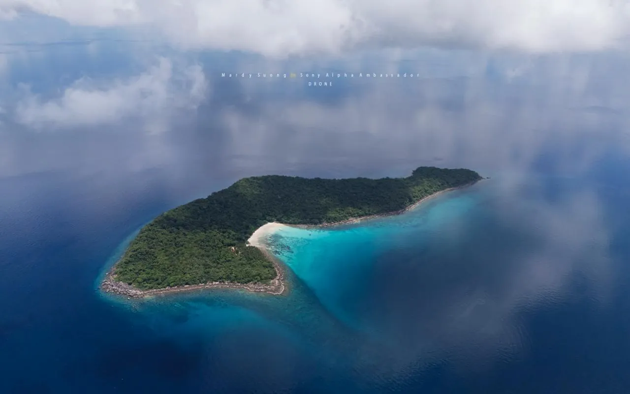 A forested island ringed by turquoise shallows, seen from above through clouds