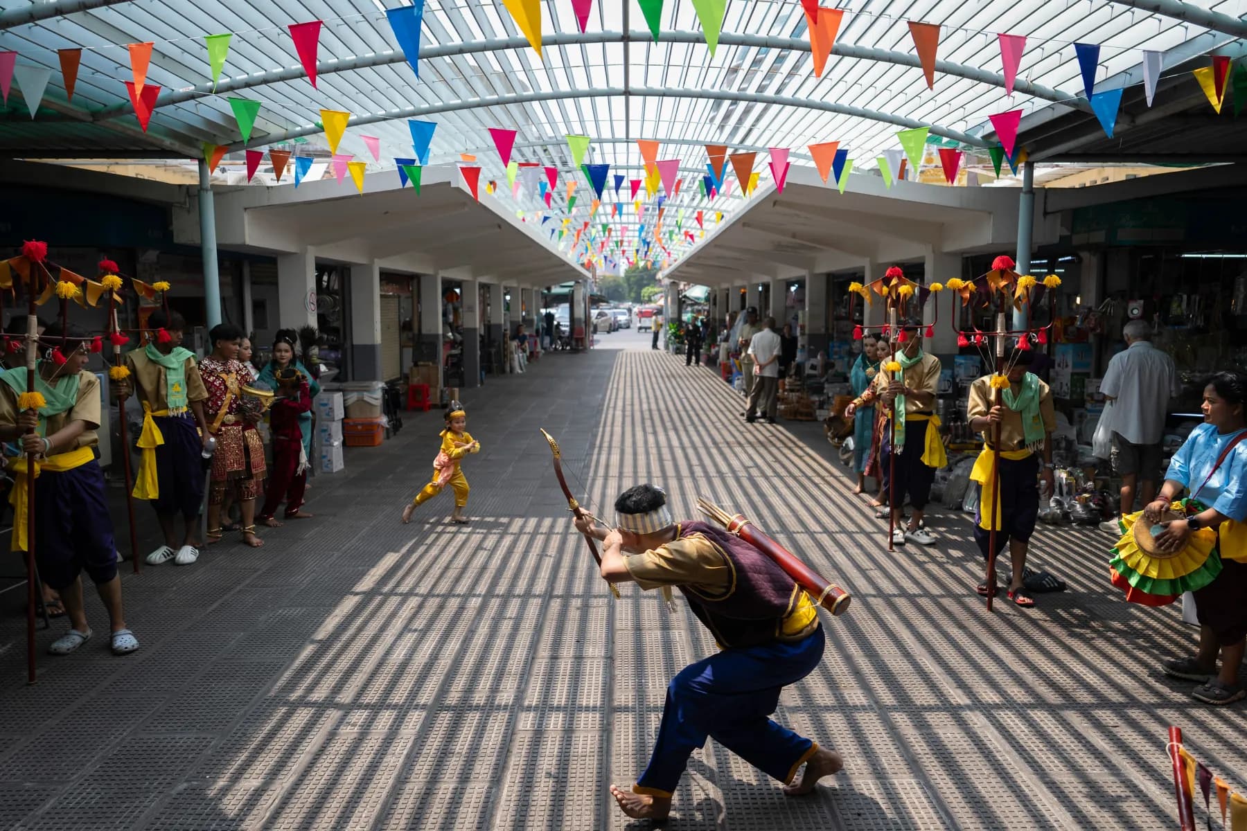 A Robam Trot dancer lunging with a staff under strings of bunting at Phnom Penh’s Central Market, a child dancing behind him