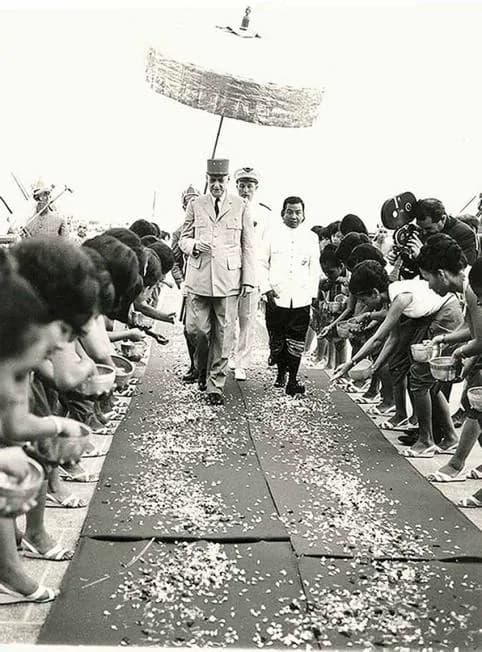 President Charles de Gaulle walking between rows of people under a ceremonial parasol