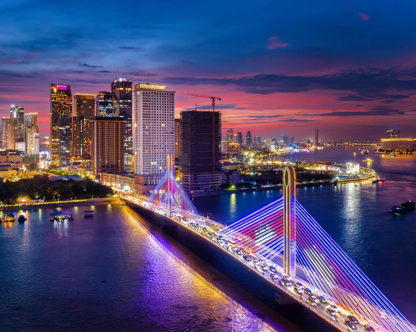 A bridge lit in colour at night, with the city’s towers behind