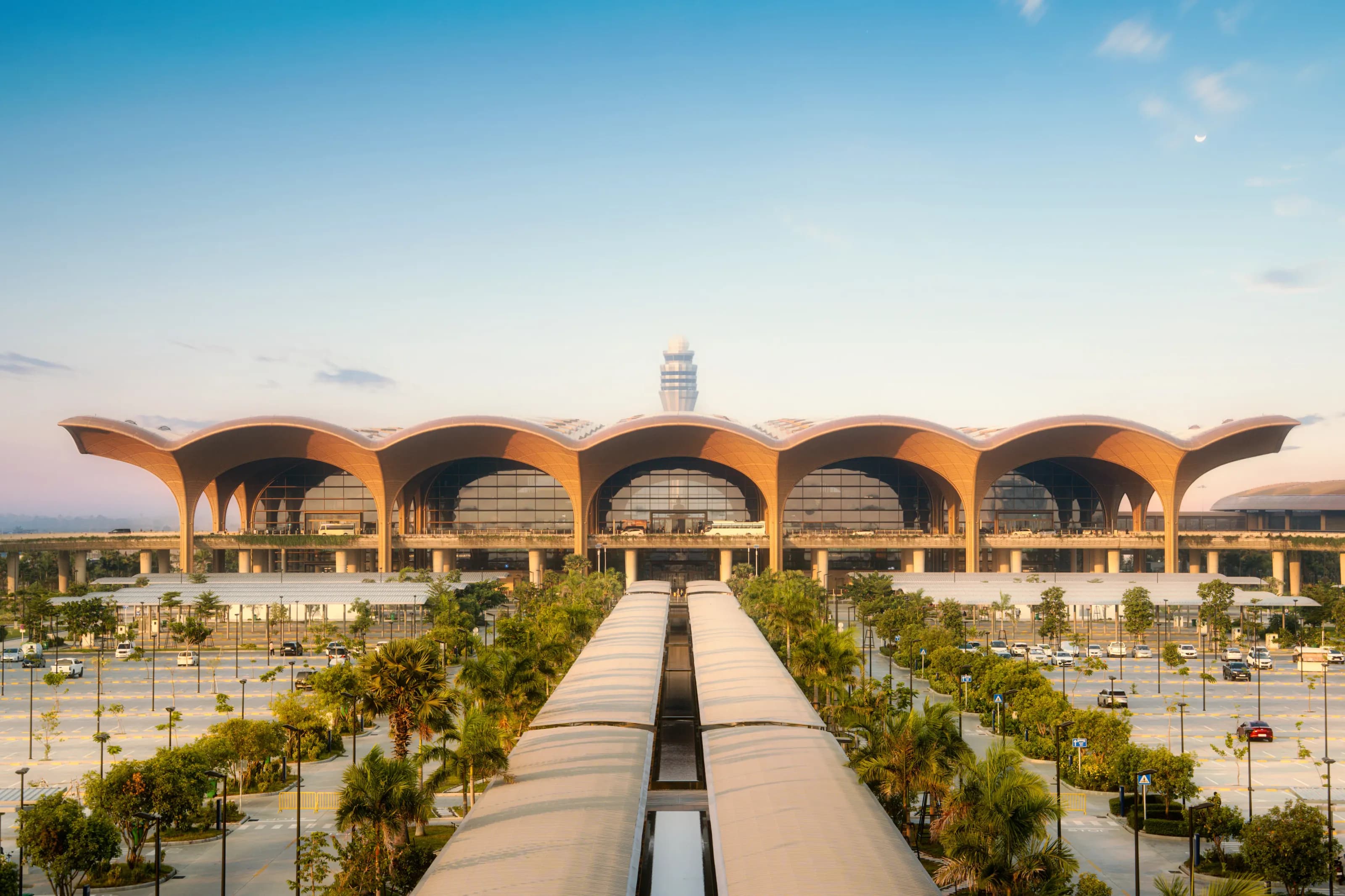 The terminal of Techo International Airport, a row of arched wooden roofs with the control tower behind, above palm-lined walkways